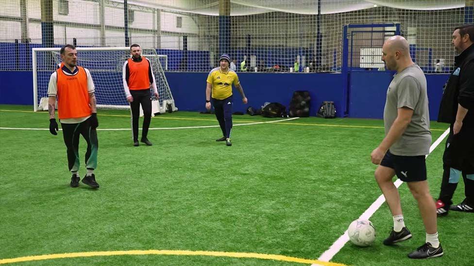 On the indoor pitch at one of the football sessions. Three men stand in front of the goal in sports wear, two with orange bibs, while another is about to kick the ball on to the pitch