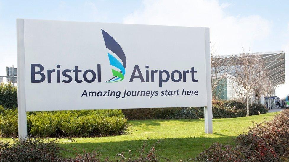 The entrance to Bristol Airport, with a large white sign saying "Bristol Airport, amazing journeys start here". The main terminal building can be seen in the background, alongside some landscaped greenery. 