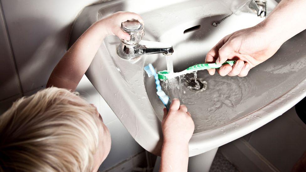 A blonde haired little boy holding his toothbrush under a bathroom tap and an adult hand doing the same