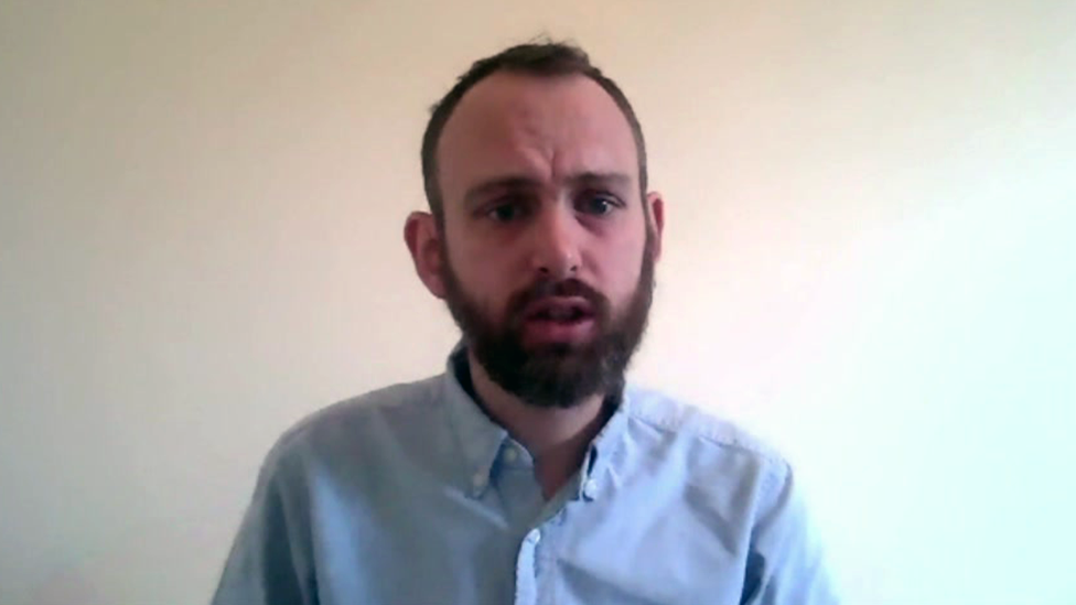 A man with a beard wearing a light blue shirt looks into the camera.