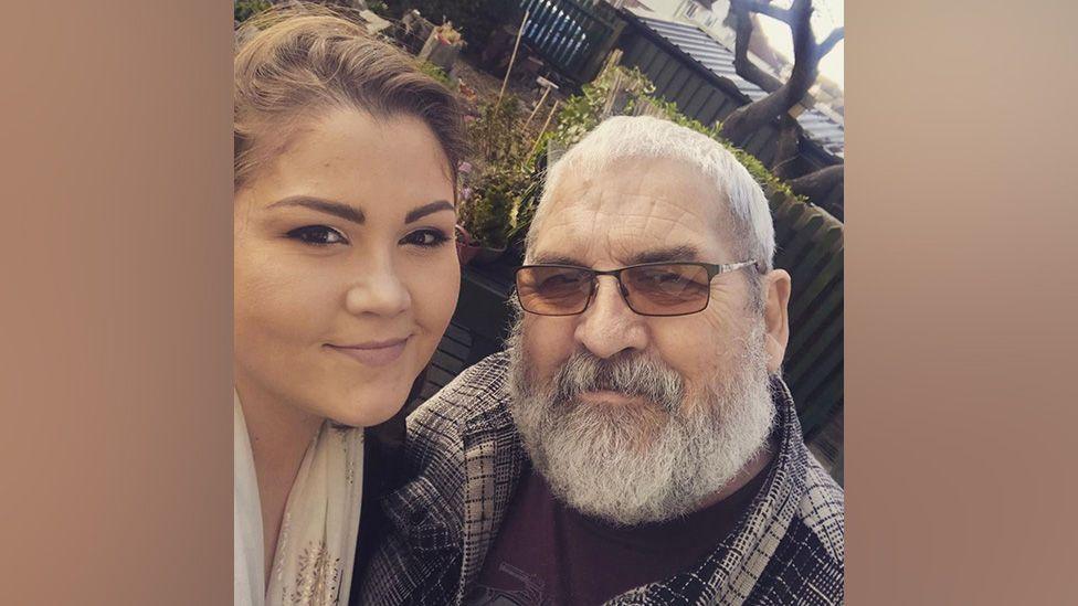 A young woman with brown hair poses next to her father, an older man with glasses and a beard, both smiling at the camera