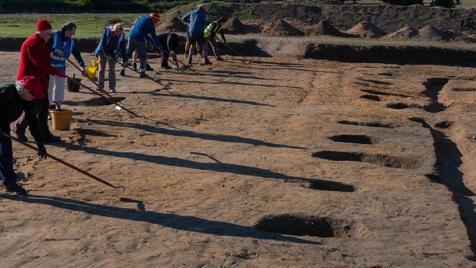 Fully excavated post holes on the east side of the hall, and volunteers cleaning the trench for post excavation photography