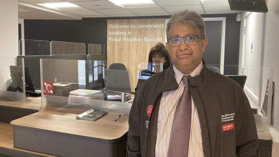 Ashwin looks into the camera in front of the banking desks with Rita behind the desk