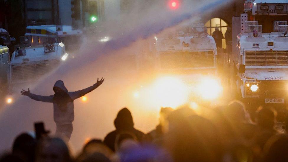 Police aim a water cannon at demonstrators. A man stretches with his arms out as a stream of water cannon is fired at him.