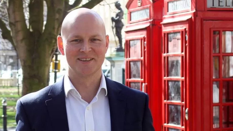 Man in a suit jacket stood in front of a red phone box in Cheltenham