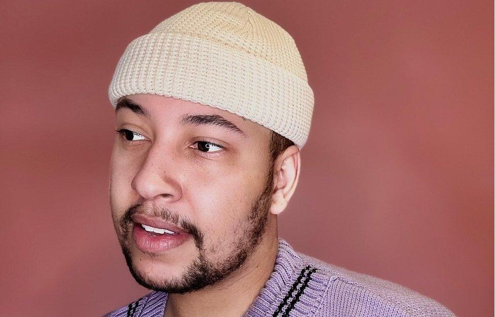 Ben Thompson poses for a studio portrait against a pale red background. He has a short beard and is wearing an off-white beanie that sits close to his head, and a pale lilac v-neck jumper. He looks thoughtful as he focuses on something outside the shot.