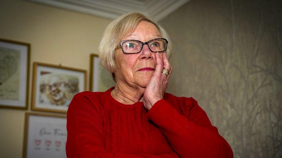 head and shoulder shot of Linda Roberts wearing a red jumper and holding her hand to her face, which looks sad