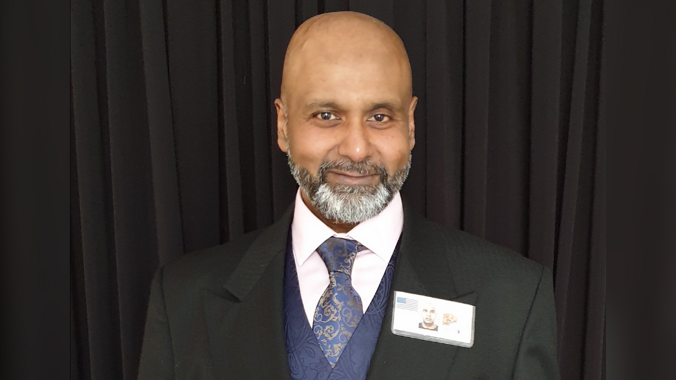 Mark Christopher, wearing a black suit jacket, mauve waistcoat, navy tie and white shirt. He is standing in front of black curtains and smiling at the camera.