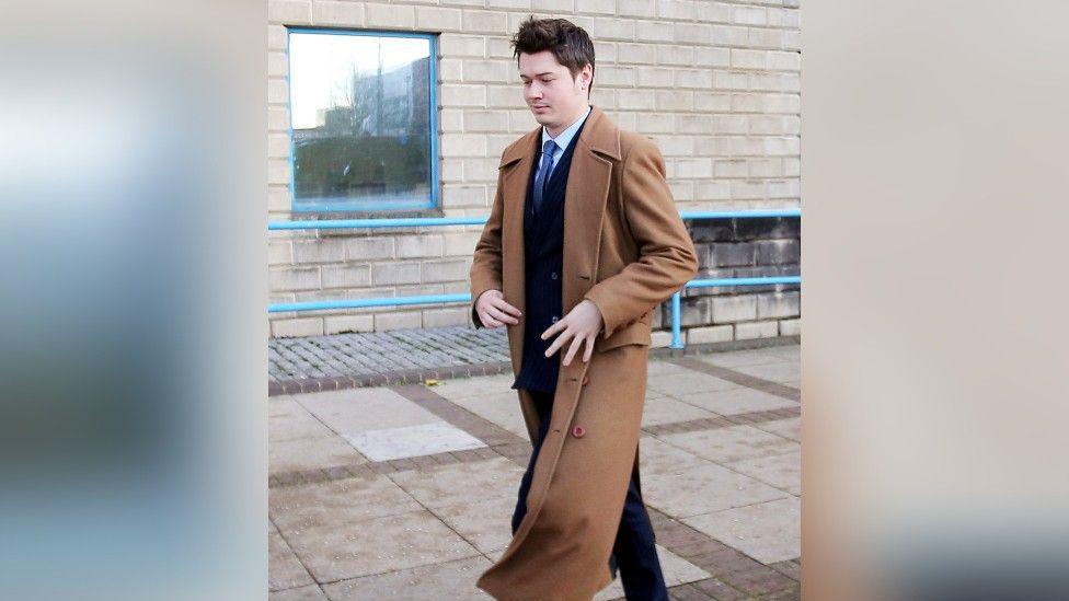 A man with brown hair, wearing a brown overcoat and a blue suit, walking down the pavement outside a court building.