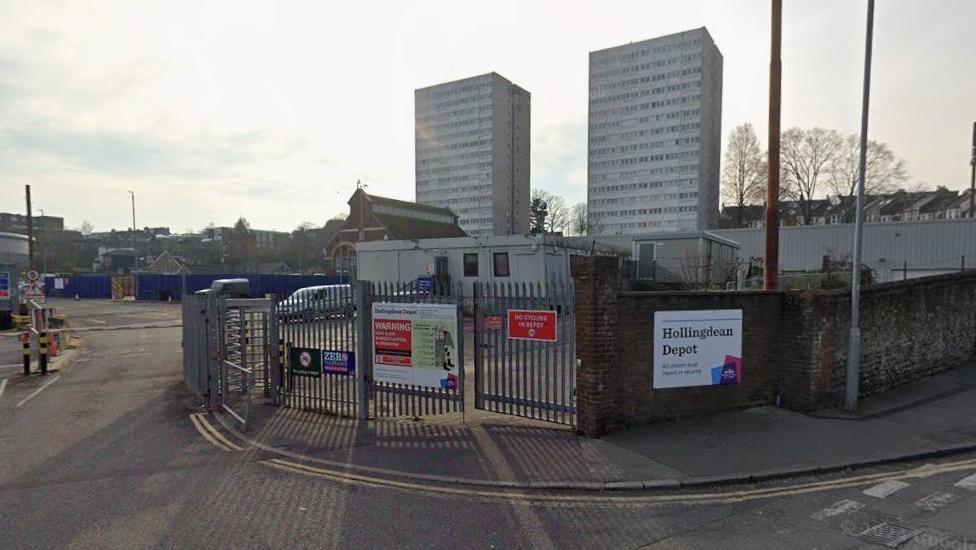 A Google StreetView image showing the entrance to the Hollingdean Depot which has large metal gates and fences and a council sign on the front with the name of the depot on it. In the background are two tower blocks.
