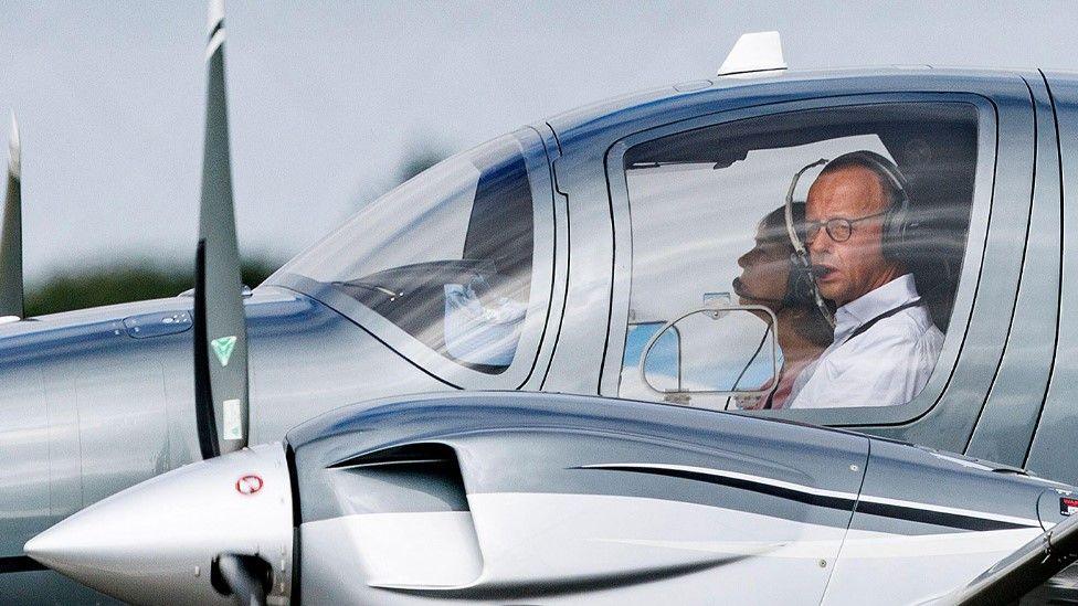 Friedrich Merz looks out of the cockpit, wearing headphones and glasses alongside another person who is not quite visible