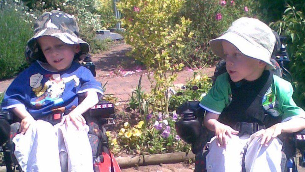 Sam and Alex sat in their wheelchairs when they were children. They are side-by-side. Both of them are wearing bucket hats and white trousers. 