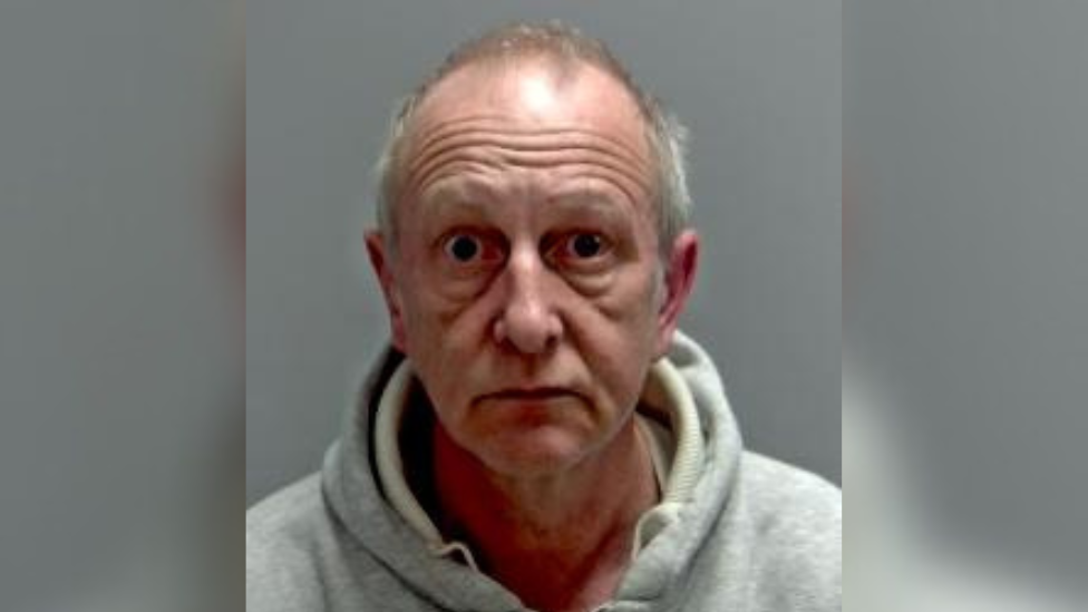 A custody mugshot taken of Martin Pearson, who is wearing a grey hoodie and has grey hair. He is wide-eyed.