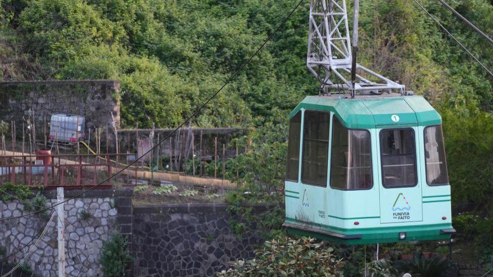 A cable car stands still in Faito, Castellammare, Italy, after the crash. It is on the right side of the picture and is light and dark green. In the background is the mountain.