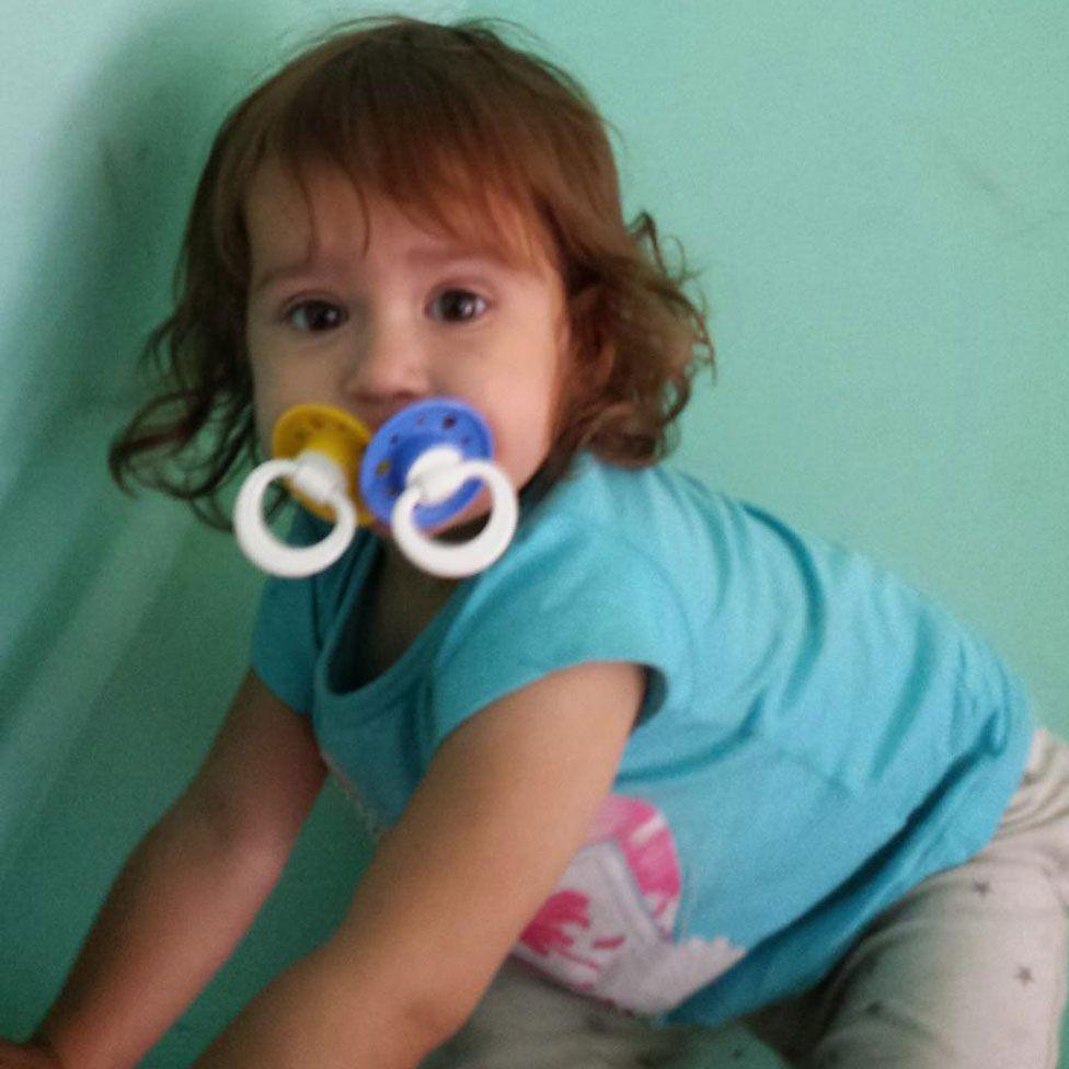 Sara Sharif pictured as a toddler with two dummies in her mouth, wearing a turquoise T-shirt and grey bottoms. 