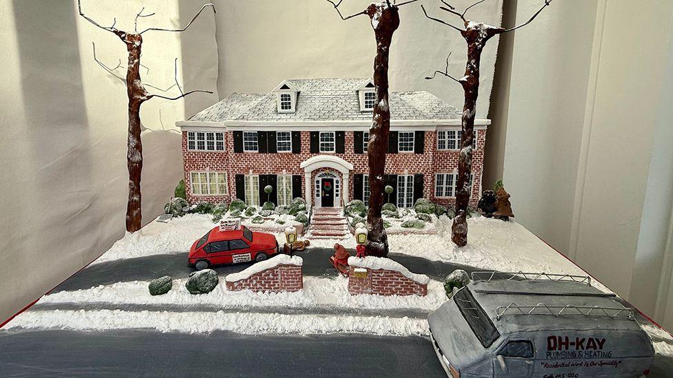 The cake in the window it shows the two-storey red brick Home Alone house with icing snow on the roof and front lawns on the driveway is a confectionary car and van in the road