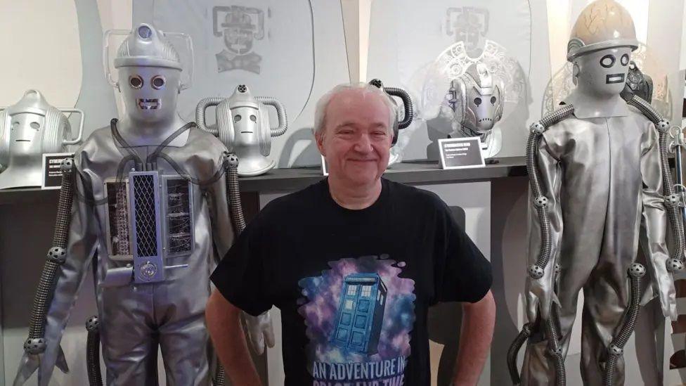 A grey-haired man smiling into the camera with his hands on his hips. He is wearing a black T-shirt with a picture of a blue police box on it, and the words "An Adventure in Space and Time". Behind him, on display as exhibits, are two Cybermen costumes - robotic-looking alien villains from the BBC science-fiction series Doctor Who.