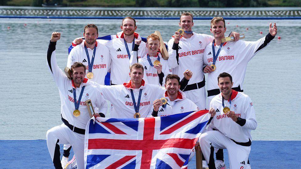 Men's eight posing for a photo with their medals by the river wearing white tracksuits