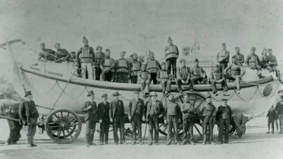 Black and white photograph dated 1865 of lifeboat sat on a wheeled structure and crew onboard and alonside the vessel.