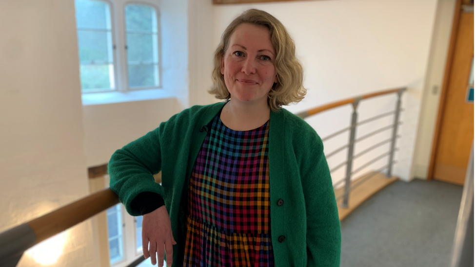 Woman in green jumper stood in a hallway with right arm on a railing