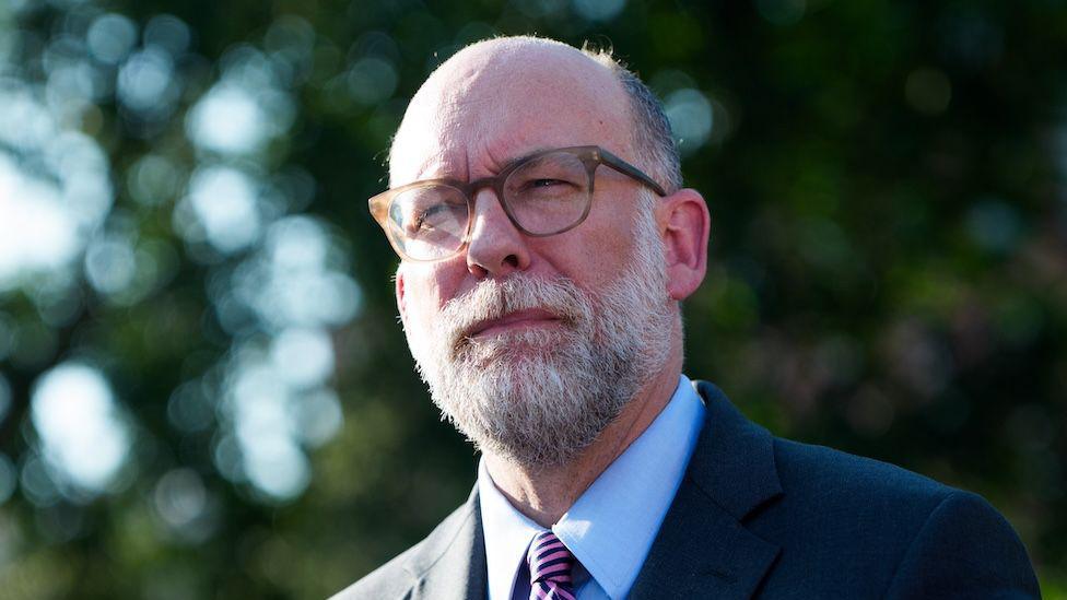 Russell Vought speaks to media outside wearing his glasses, a striped tie, button-down shirt and dark suit jacket