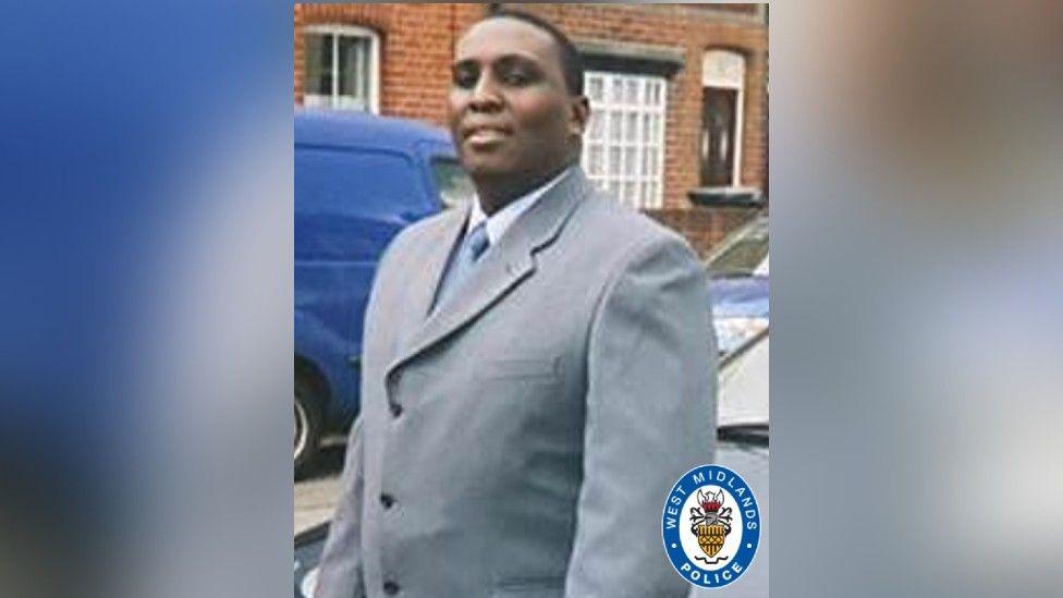 A man with short-cropped black hair is wearing a light grey suit jacket over a white shirt and pale blue tie. A West Midlands Police logo has been superimposed in the corner of the picture.