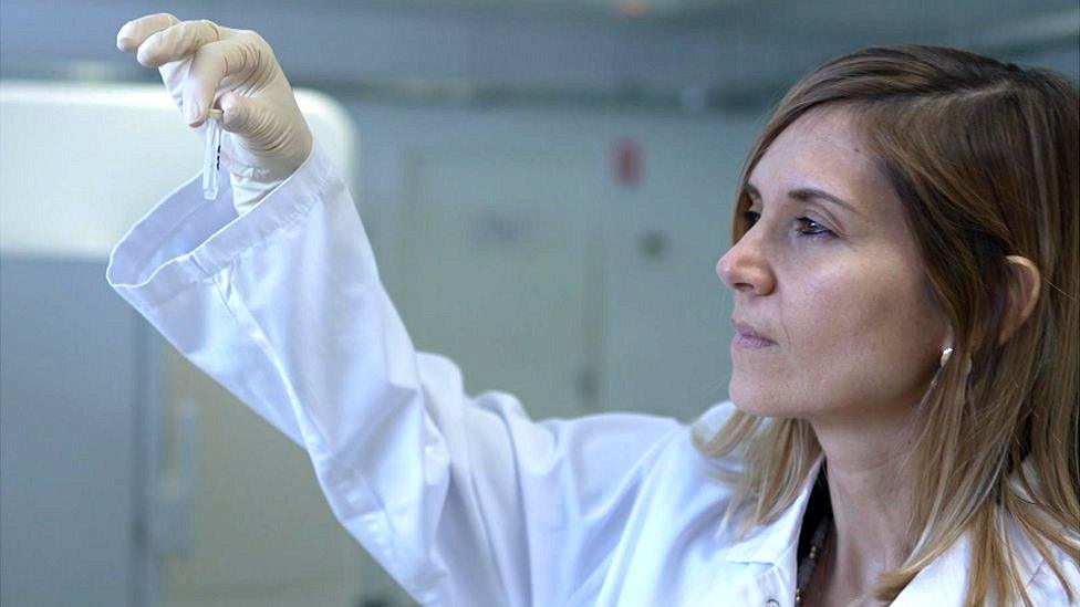 Dr Mathilde Touvier is wearing a white lab coat, with her right hand she is holding up a small test tube and looking at it