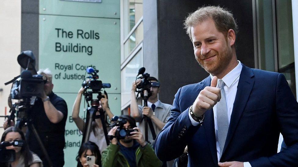 Prince Harry, wearing a blue suit, blue shirt and tie, grins and gives a thumbs up to the camera, as he walks past a sign for the Royal Courts of Justice. Six photographers and camera operators with cameras are in the background