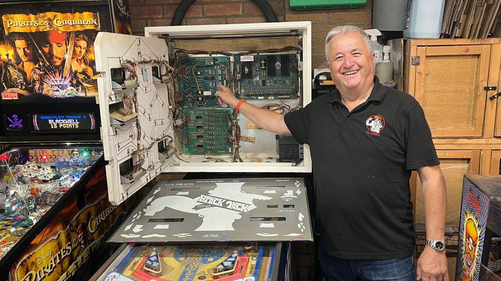 Mark Squires with short white hair, wearing a grey polo shirt, smiling at the camera, holding up his right hand and pointing to wires and connections inside a pinball machine