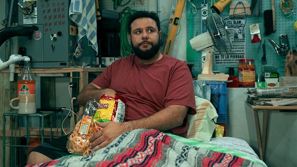 Mo in the show, wearing a red t-shirt, sitting in bed and eating crisps. Around him are tools and a fan as he looks off into the distance.