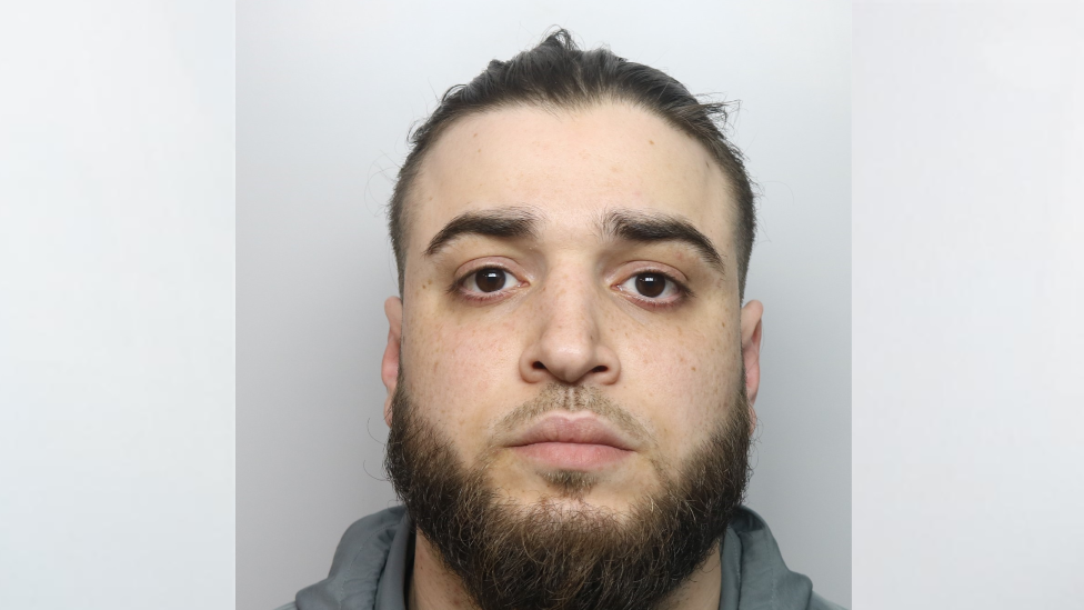 A mugshot of Hamze Almallah, who has dark hair tied back and a brown beard. He is emotionless as he stares ahead 