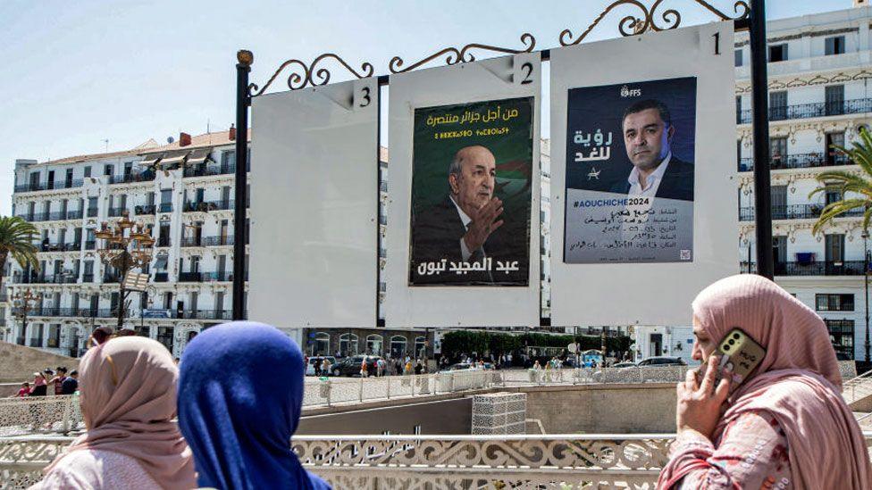 Women walk past a notice board depicting posters for presidential election candidates incumbent President Abdelmajid Tebboune (L) and the Socialist Forces Front's Youcef Aouchiche, in the centre of Algiers - 5 September 2024