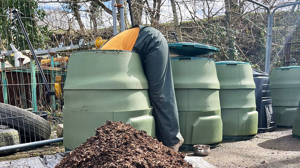Somebody wearing an orange t-shirt and green work overalls is bent over leaning a long way into a green composting or storage bin. There is a pile of compost in front of the bin. Three others stand to the right of it. Behind is movable metal fencing with tyres and other industrial type equipment behind it, and a tree and scrub covered embankment.