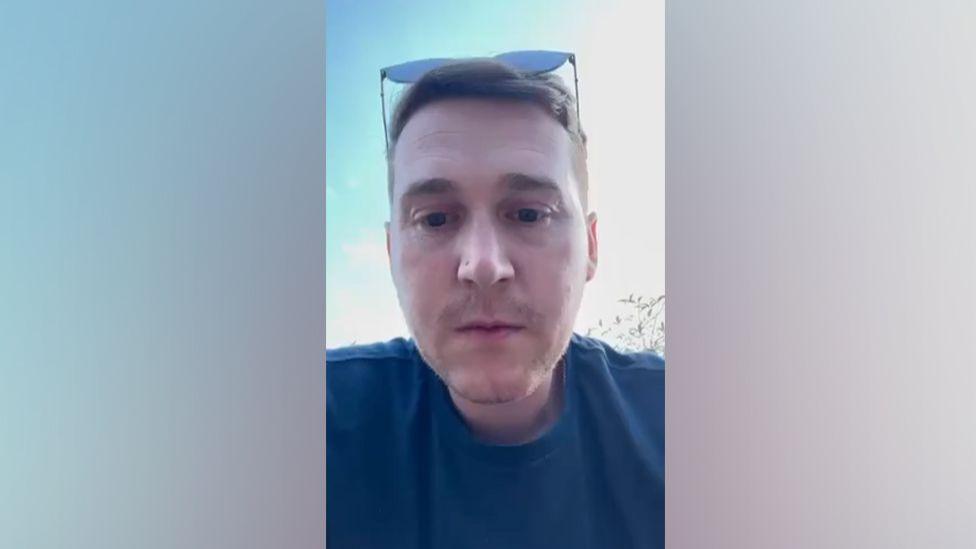 Ben has short brown hair and a pair of sunglasses are resting on the top of his head. He is wearing a blue t-shirt.