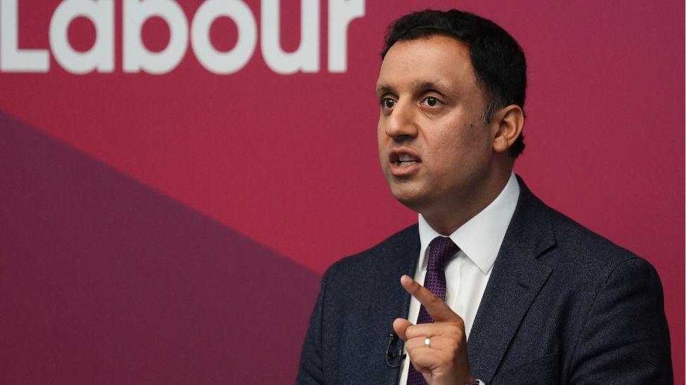 Anas Sarwar, a man with dark hair, speaks while standing in front of a red Labour sign. He is wearing a dark suit, white shirt and purple tie, and pointing with his left hand.