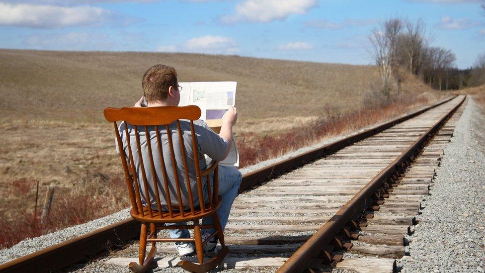 A man reads newspaper whilst sitting on a rocking chair placed on a railroad track