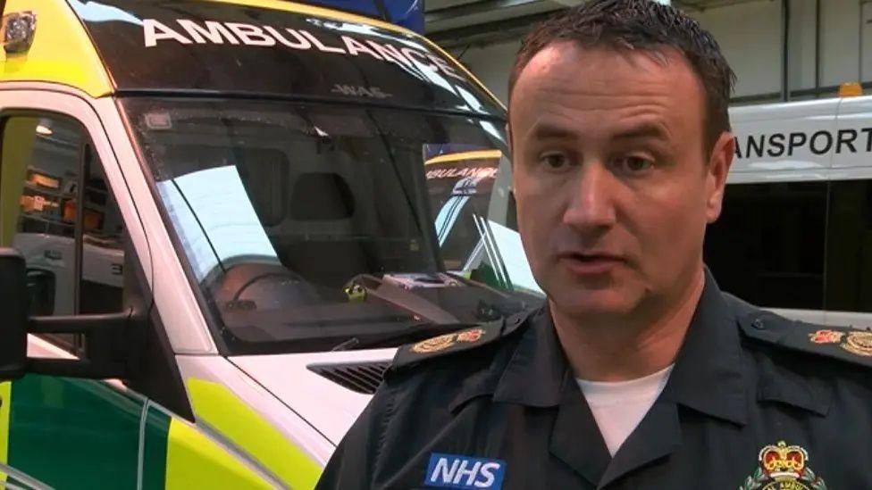 Mark Ainsworth in NHS ambulance uniform standing in front of a green and yellow paramedics vehicle 