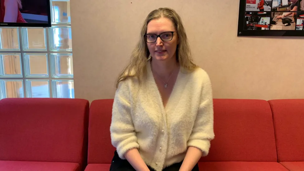 Catherine Flanagan wearing glasses and a cream cardigan sat on a red sofa in the BBC building