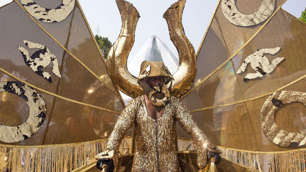 A man in a gold sequin outfit with a giant manilla behind him holding up gold wings takes part in a carnival in Calabar, Nigeria