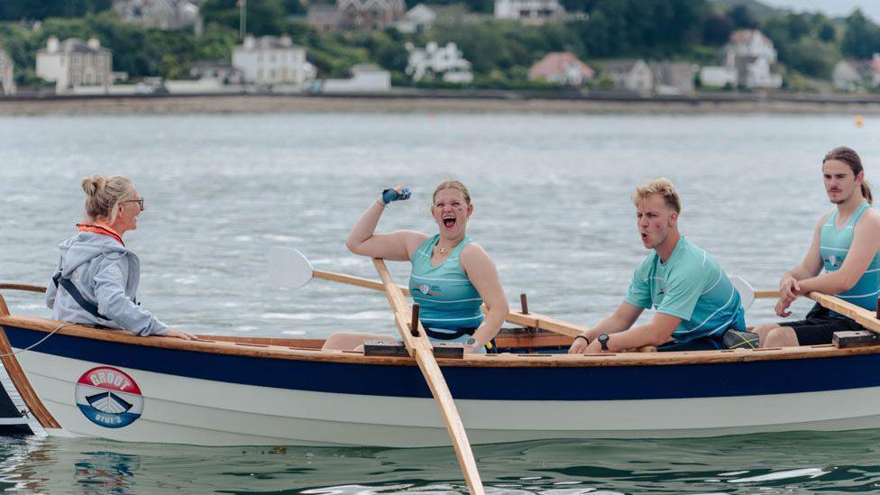 A female rower in a light blue/turquoise top punches the air while the rest of her crew look tired around her