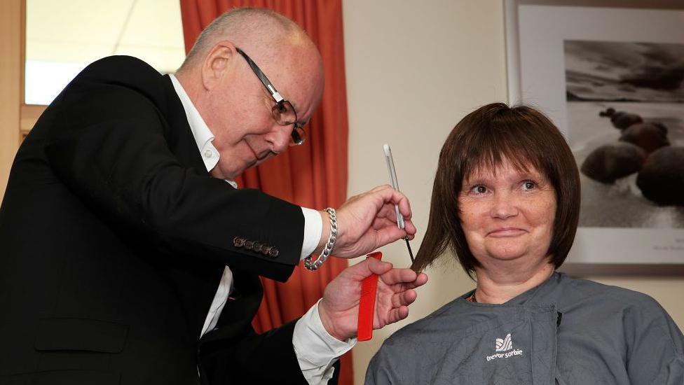 Trevor Sorbie styling the wig of a cancer patient. He is wearing a black jacket and white shirt and has a bald head and glasses. He is using scissors to style a brown wig on the head of a woman who is wearing a grey Trevor Sorbie hairdressing cape. He is also holding some red scissors and has a chunky bracelet on his wrist. There is a red curtain and a black and white framed picture in the background.