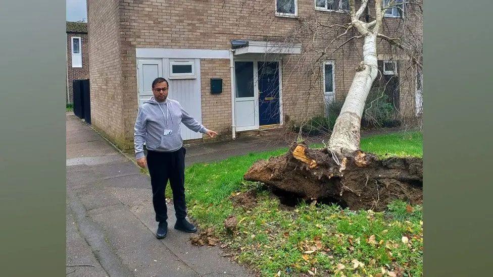 A man stood next to a tree in the front garden of his house. He is pointing at the tree