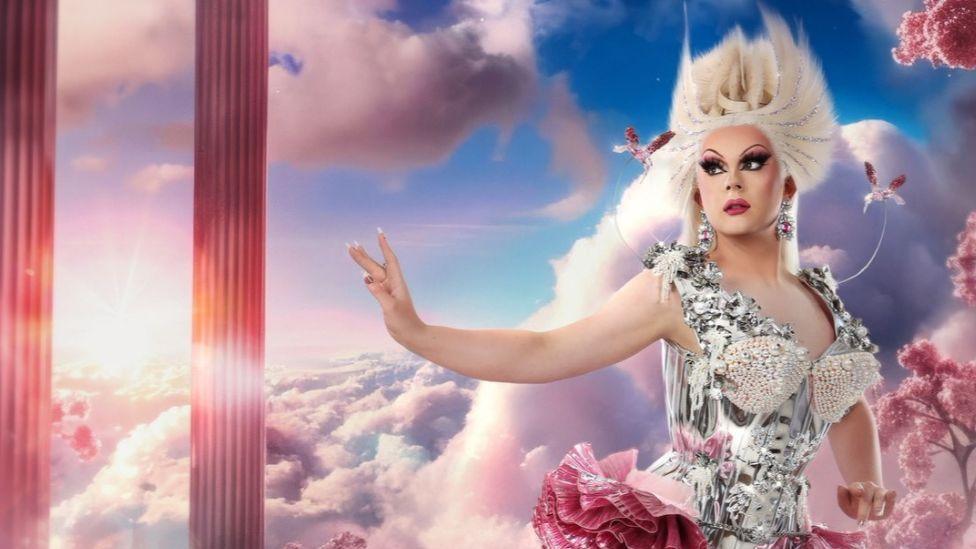  Bonnie Ann Clyde. She is wearing a dress with a silver corset and a pink ruffled skirt with a leg slit. She is also wearing a blonde wig which is styled to stick up. She is standing in front of a background of blue skies, green grass and large pink crystals. 