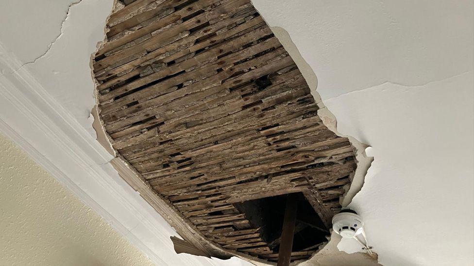 A damaged ceiling showing bare wood boards where the plaster has come off and a square hole