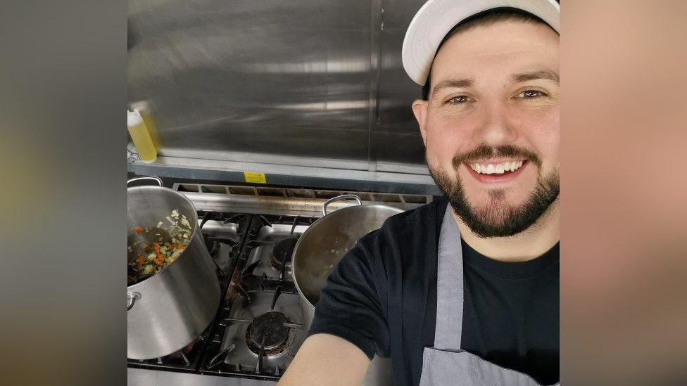 A man with a white hat and dark brown facial hair is taking a selfie in front of a hob with two silver pans on it