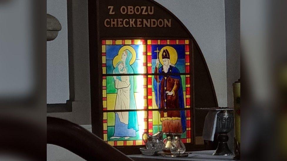 A colourful stained glass window propped up inside a church with the word "Checkendon" above it.
