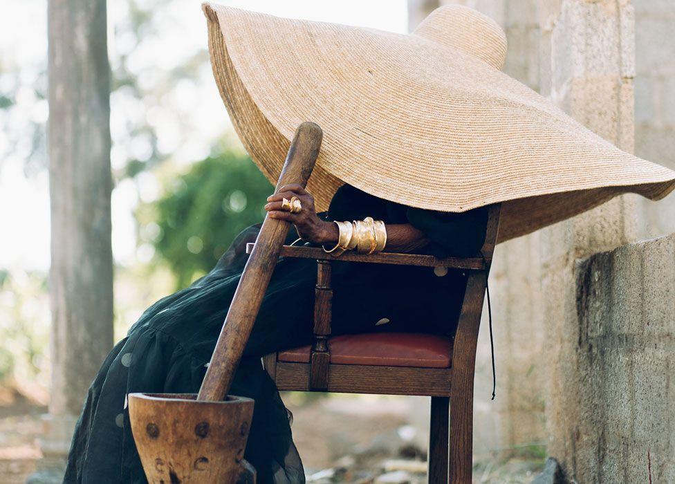 A side view of Margret Chola in a long black-netted dress sitting outside on a wooden chair wearing an enormous straw hat that obscures her profile. Her hand, on which she has a big gold ring and big gold bracelets, holds a long wooden stick that is used to pound grains and stands in a wooden mortar