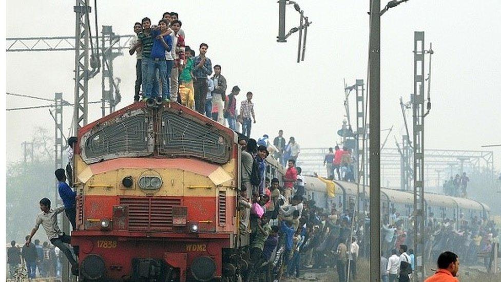 passengers stand on top of a moving train, hang onto the sides and run along beside it, trying to get on