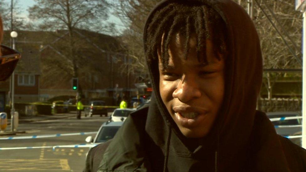 Eric standing at the scene of the incident wearing a black hoodie. Blue police tape can be seen blocking the road behind him