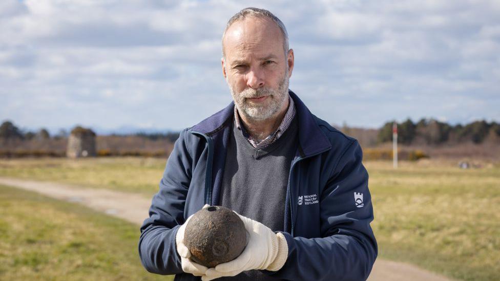 A man standing on Culloden battlefield holding the unexploded shell. He is wearing white gloves and a blue NTS fleece. 